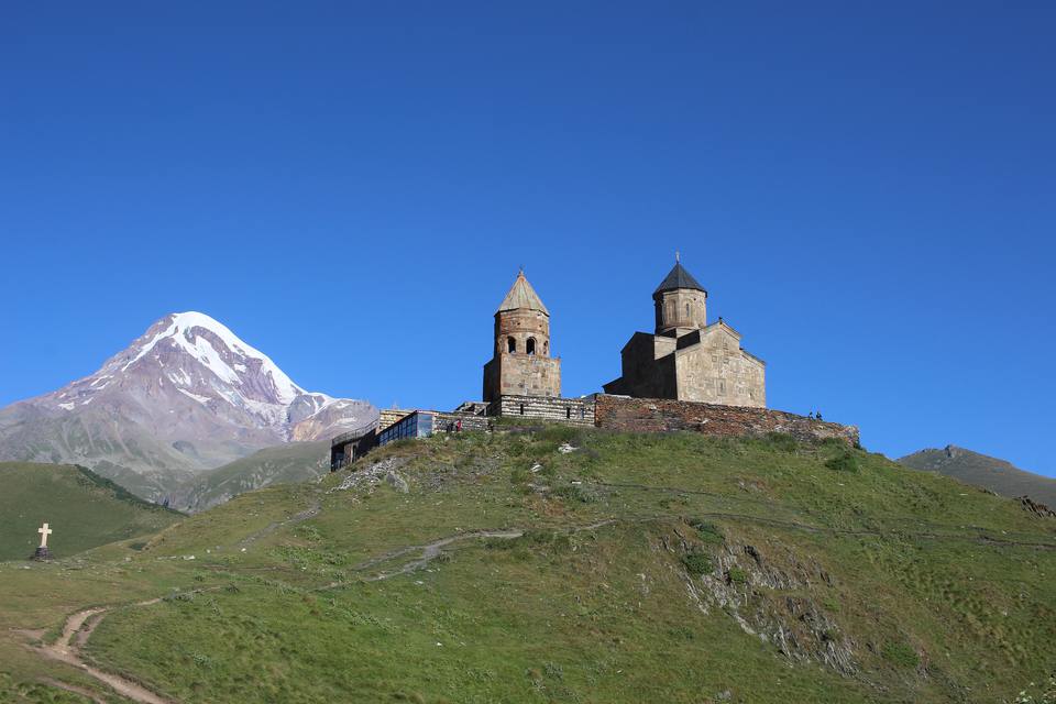 Gergeti Trinity Church and its bell tower on a green ridge above Stepantsminda, with the snow-capped summit of Mount Kazbek behind, under a cloudless blue sky