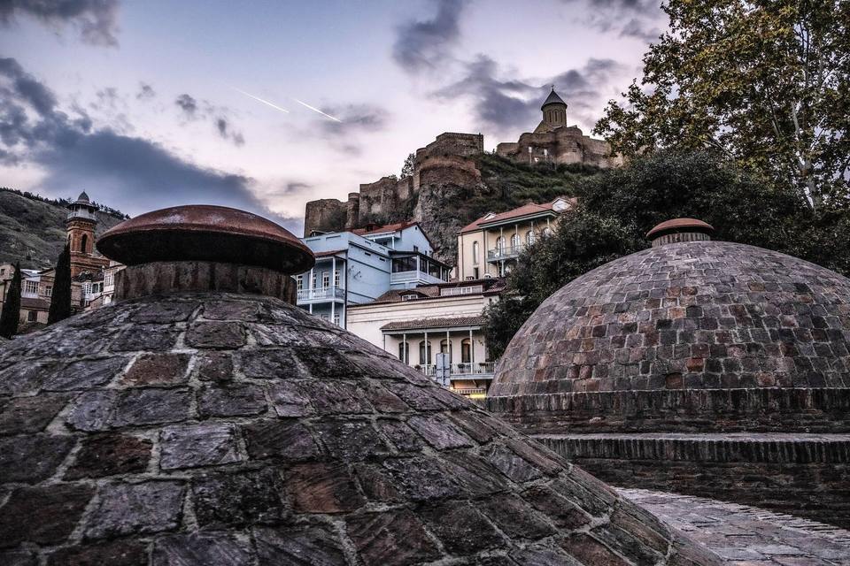 Brick domes of the Abanotubani sulphur baths at dusk, with balconied old-town houses and Narikala fortress on the hill behind, Tbilisi, Georgia.
